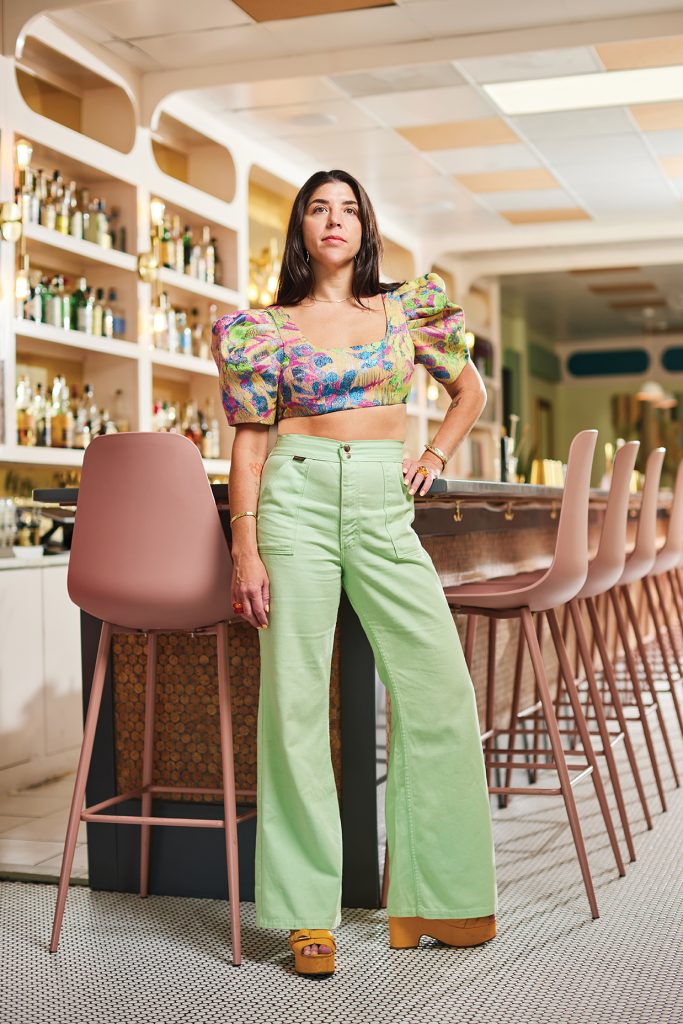 Woman in colorful crop top and mint green pants standing at a bar