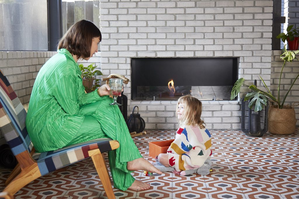 A woman in a bright green outfit sitting in a colorful chair talking to a young girl sitting on a patterned tile floor in front of a white brick fireplace