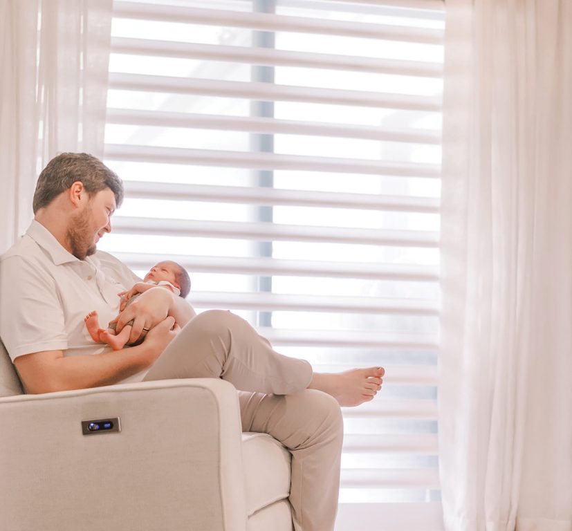 A person sitting in a recliner and holding a baby in front of a window
