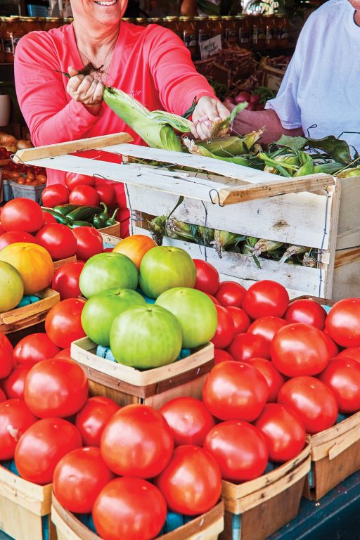 Rows of baskets holding red, ripe tomatoes and a woman shucking corn in the background
