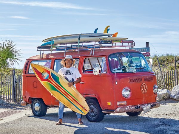 A smiling man holding a surfboard and standing in front of an old, red Volkswagen bus