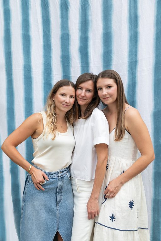 three young women standing close together in front of a blue and white striped tapestry