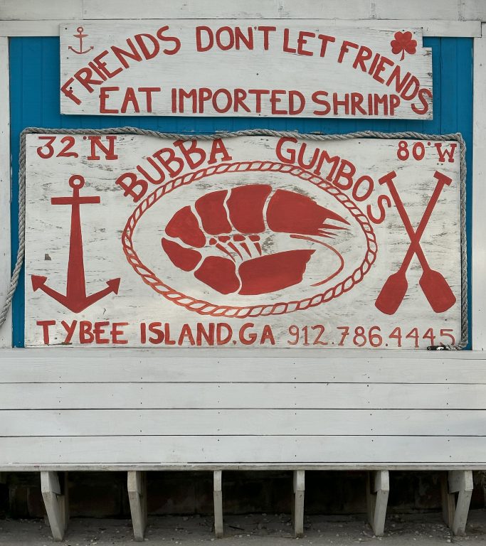 A white bench under a hand-painted wooden sign that says "Friends don't let friends eat imported shrimp."