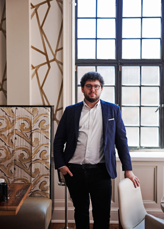 Man with glasses, curly hair and a beard standing with his hand in his pocket in front of a big window