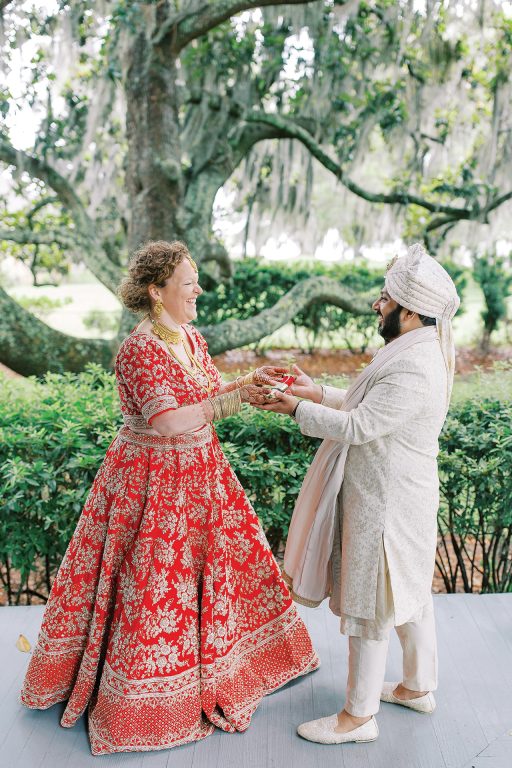A bride and groom in traditional Indian wedding attire holding hands and smiling at each other