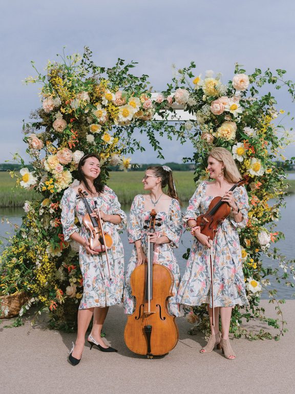 Three women holding violins and a cello in front of an archway of flowers