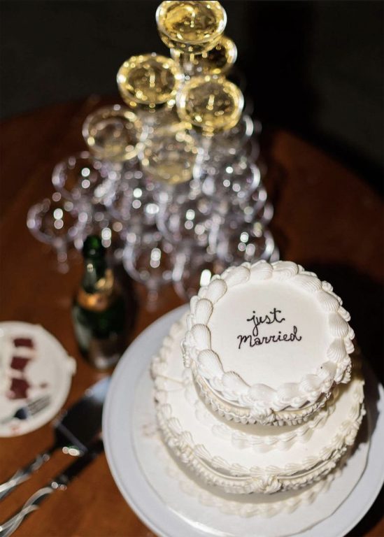 A white wedding cake with "Just Married" written on the top tier, next to a tower of champagne glasses