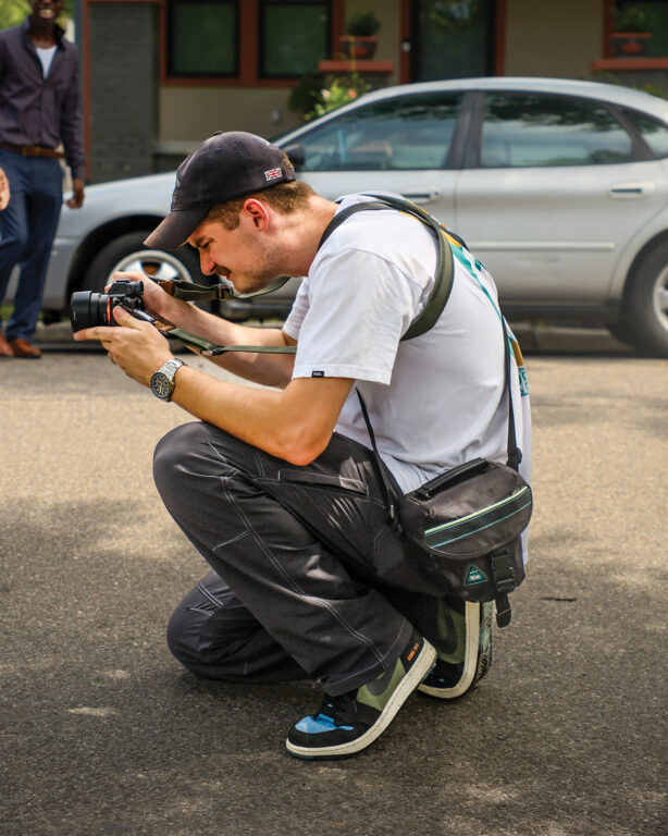 A man in a baseball cap kneeling in the street and taking a photo