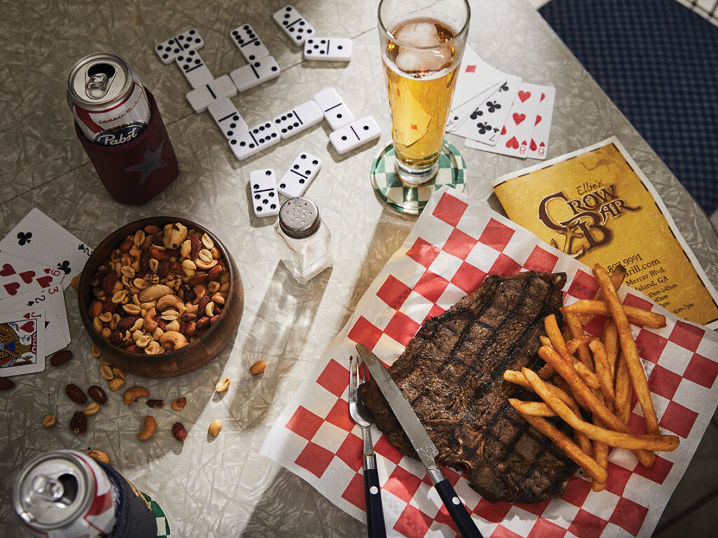 A beer, a plate of steak and fries, a bowl of mixed nuts, dominos and playing cards on a table