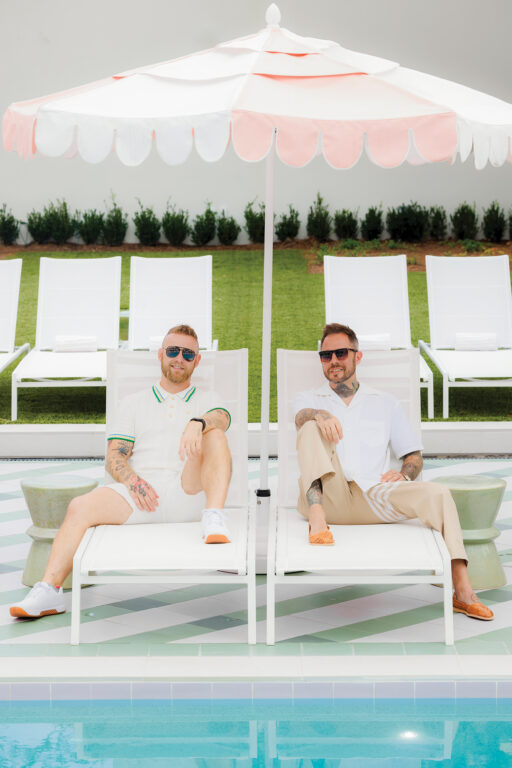 two men sitting in lounge chairs under an umbrella near a pool