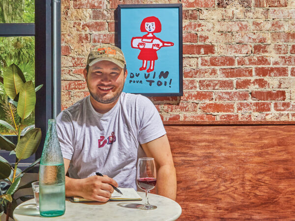 A man in a baseball cap sitting at a table with a pen, paper and glass of wine