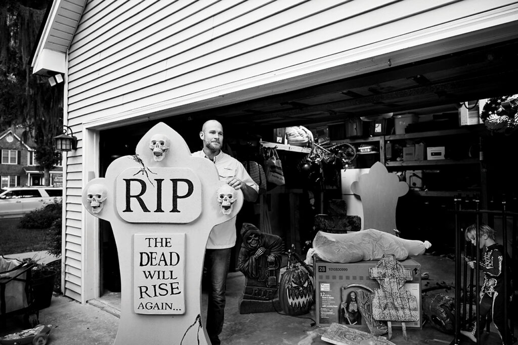 A man standing in his driveway holding a halloween decoration that says RIP