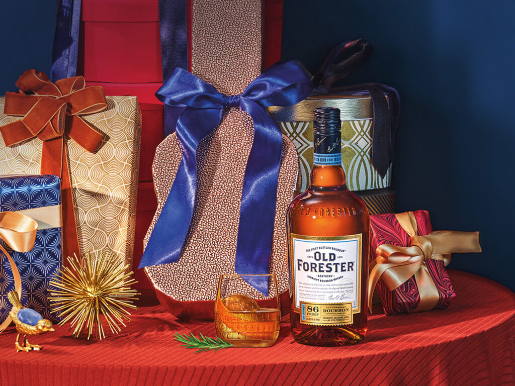 A glass and a bottle of Old Forester whiskey on a table with wrapped gifts