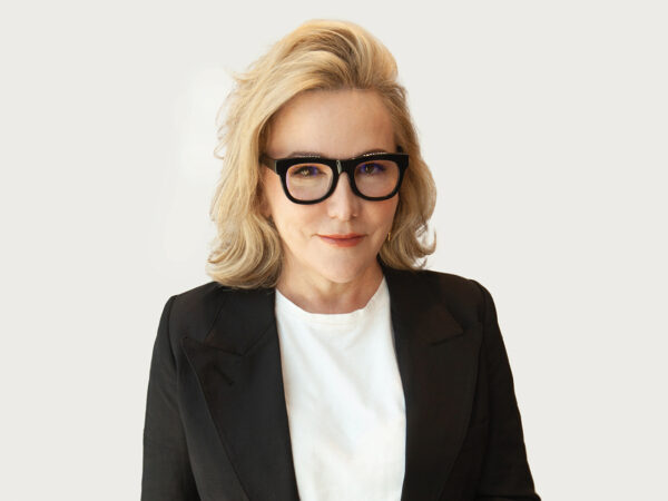 Middle-aged blonde woman wearing glasses with thick black frames, a white shirt and black blazer