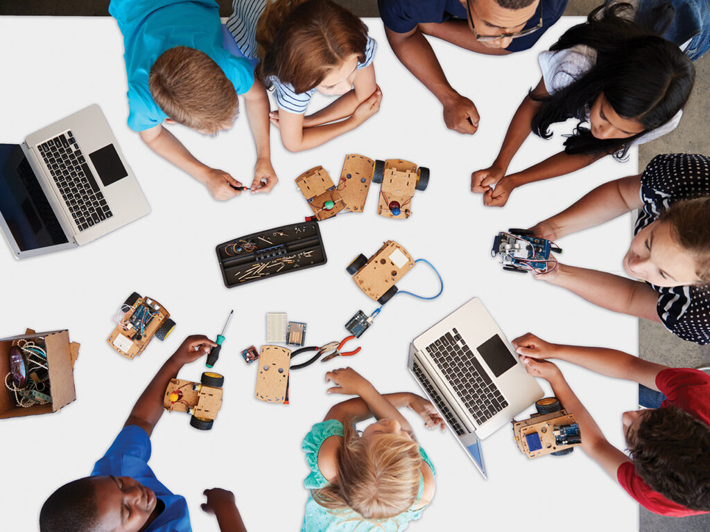 elementary school children and teacher gathered around a table with computers and school supplies