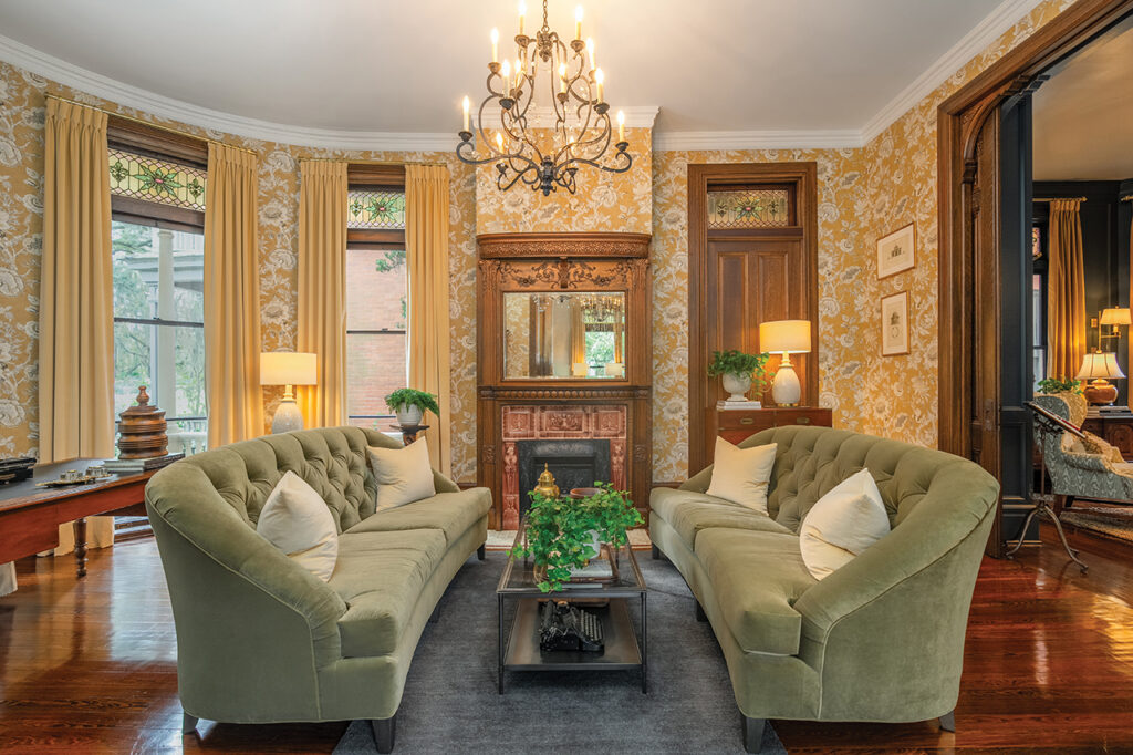 Living room in a Victorian house with gold-colored flowered wallpaper, green couches and original fireplace