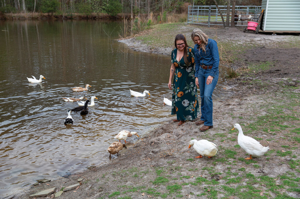 tow women standing at the edge of a pond with a group of ducks