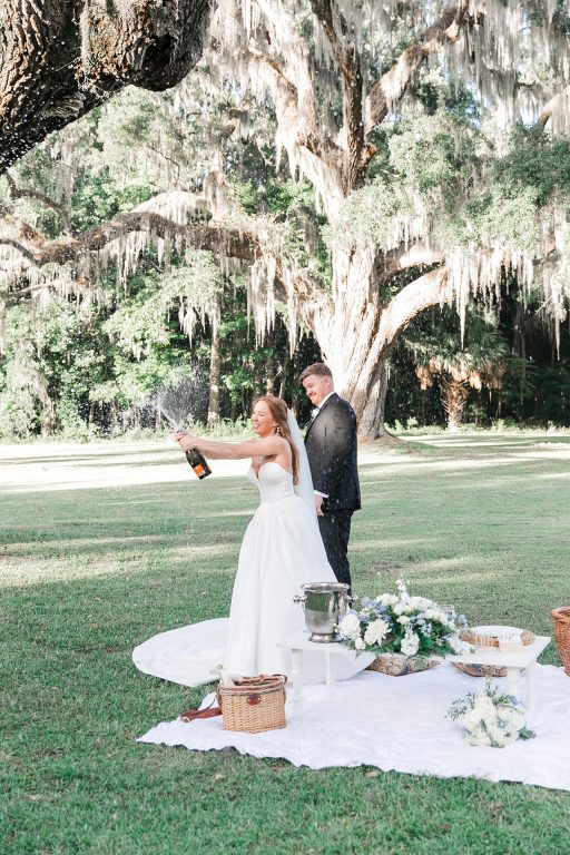 A bride and groom uncorking a bottle of champagne at a picnic