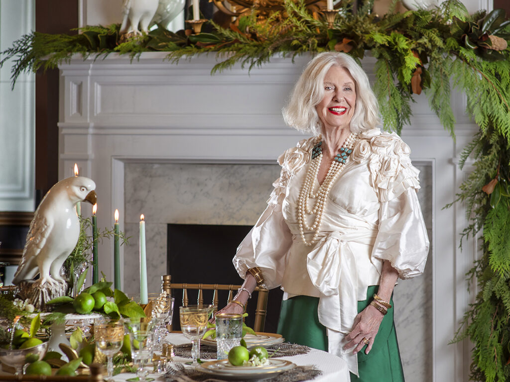 A stylish woman wearing a white satin top and emerald green satin skirt standing next to a table setting and in front of a huge fireplace adorned with greenery