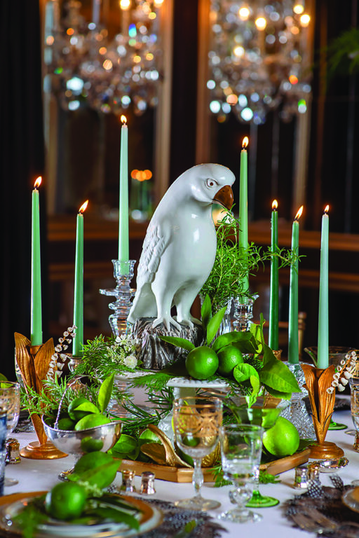 A fancy table setting with green candles, limes, rosemary sprigs, and a ceramic statue of a hawk as the centerpiece