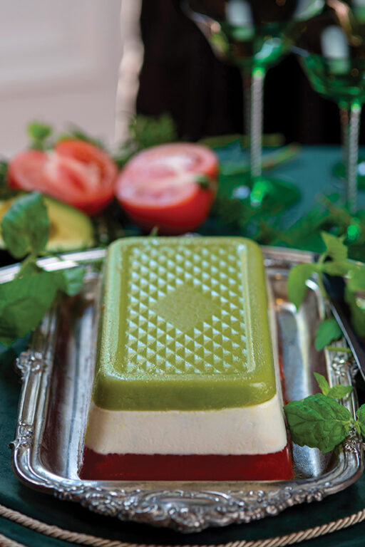 A salad mold with red tomato layer on the bottom, a white cream cheese layer in the middle and a green avocado layer on top, sitting on a silver serving platter.