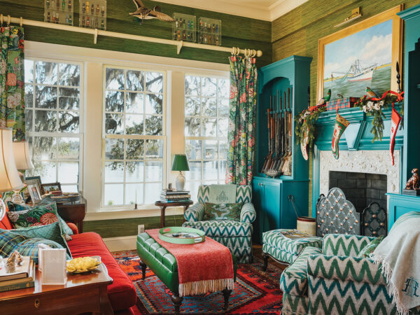 A living room with a oyster shell fireplace, teal gun shelves, green-and-white chevron armchairs, a red couch, and a green leather footstool.