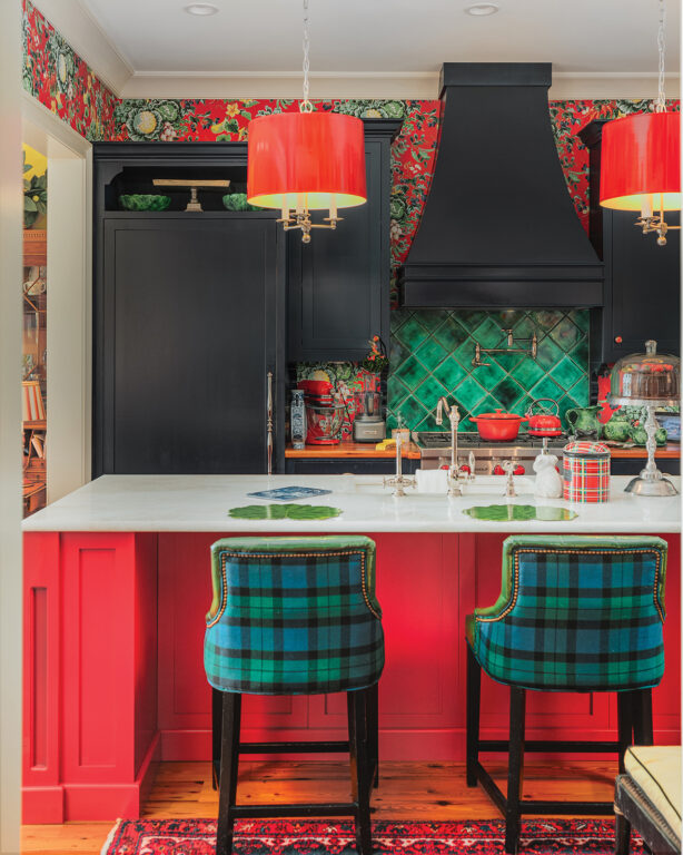 A kitchen with a red island with white marble countertop, a green tile backsplash, red lampshades, green-and-blue plaid bar stools, and black cabinets
