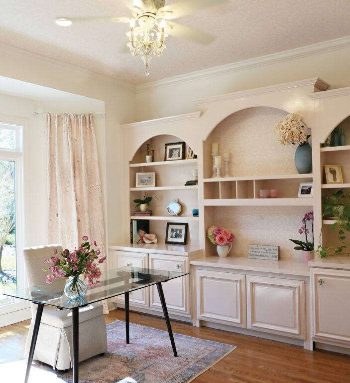 a home office with light pink built-in shelves and a glass-top desk