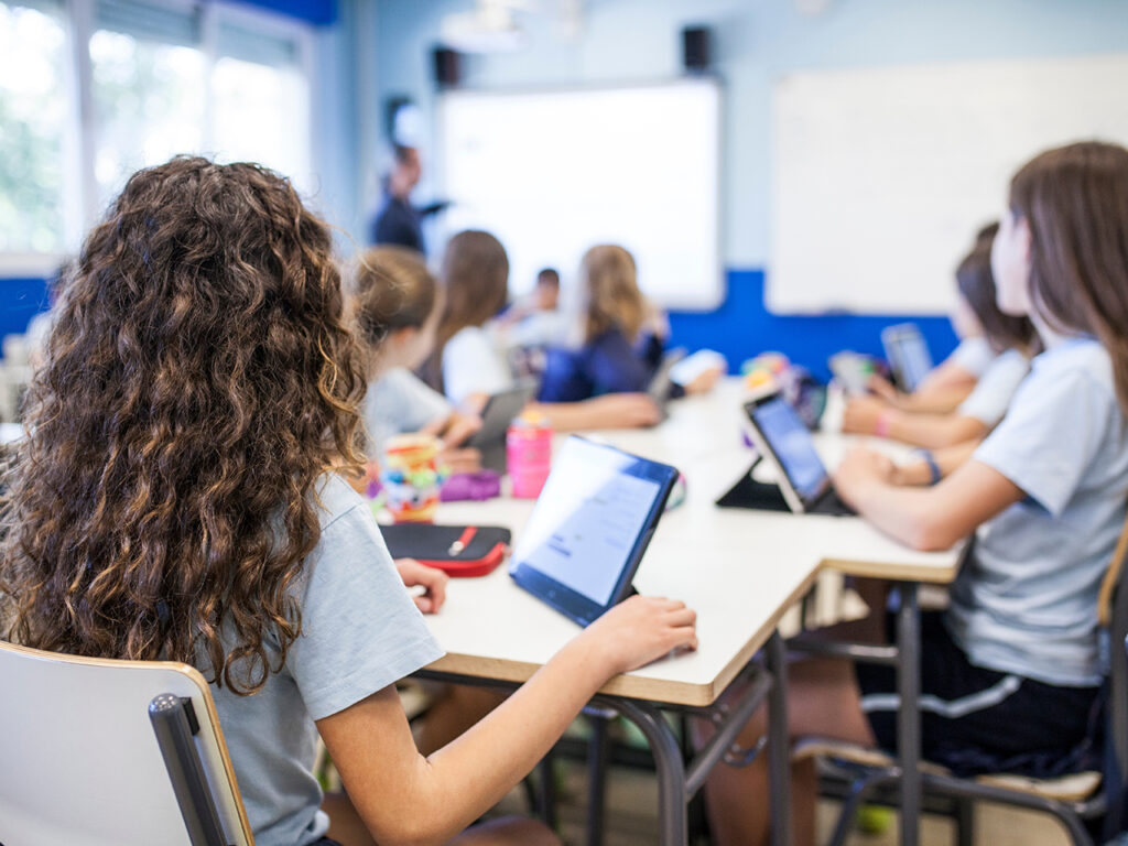 girl with brown curly hair studies in class with her tablet next to her classmates while they listen to the teacher
